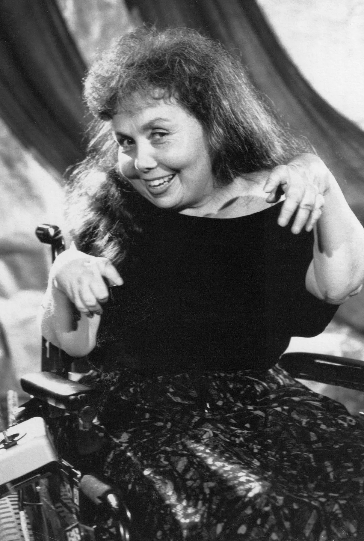 Black and white photograph of Cheryl Marie Wade in her power wheelchair. She has long, full, dark hair and smiles playfully at the camera, holding her hands up in front of her as if showing them off. She wears a black top and a patterned skirt, with draped fabric behind her.